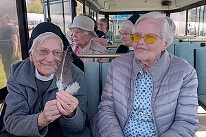 Zwei ältere Frauen sitzen nebeneinander in einem Bus, eine hält eine weiße Feder in der Hand