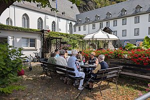 Gruppe älterer Menschen sitzt an einem Tisch auf einer Bank im Innenhof eines historischen Gebäudes mit weißer Fassade und roten Blumen