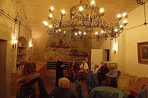 Menschen sitzen und stehen in einem historischen Raum mit Steinwand und großem Kronleuchter