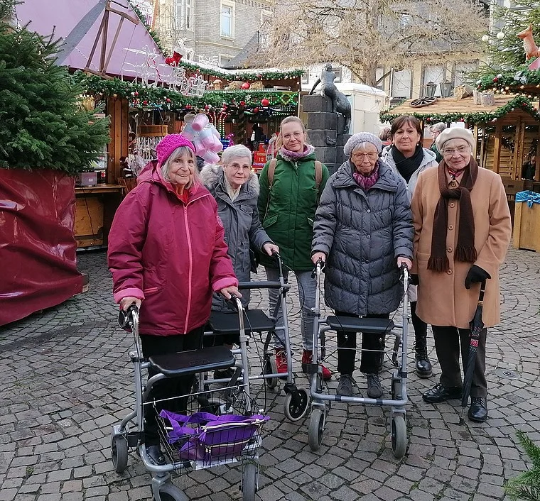 Gruppe älterer Menschen mit Rollatoren auf gepflastertem Platz vor weihnachtlich geschmückten Marktständen