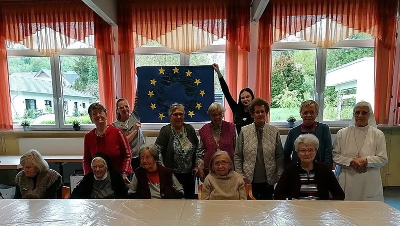 Gruppe älterer Menschen in einem Raum mit großen Fenstern, zwei Personen halten eine Europaflagge mit gelben Sternen auf blauem Grund hoch