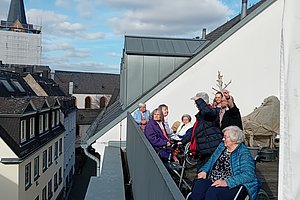Gruppe älterer Menschen sitzt und steht auf einer Dachterrasse mit Blick auf umliegende Gebäude