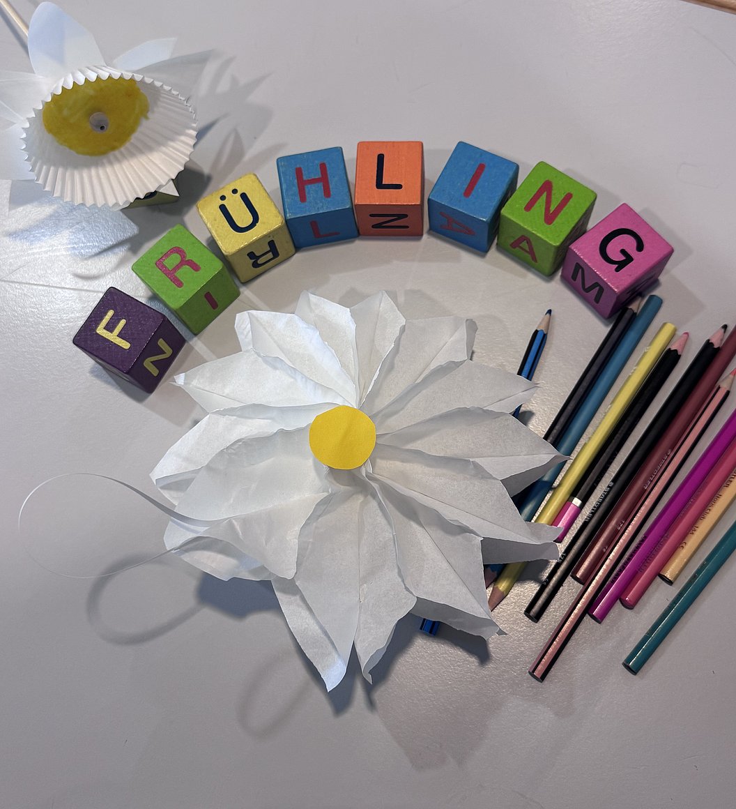 Bunte Holzbausteine mit den Buchstaben FRÜHLING neben zwei Papierblumen und mehreren Buntstiften auf hellem Untergrund