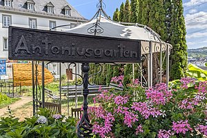 Schwarzes Metallschild mit der Aufschrift 'Antoniusgarten' vor einem Pavillon und blühenden pinken Geranien in einem Garten