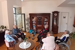 Fünf ältere Menschen sitzen in einem Wohnzimmer in einem Stuhlkreis, zwei davon im Rollstuhl, mit einem runden Tisch und einer Pflanze in der Mitte