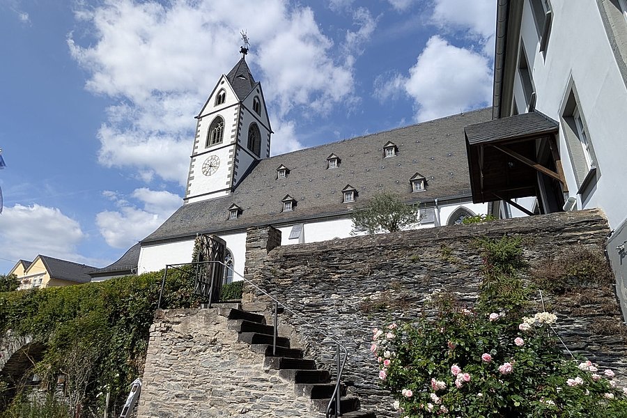 Kirchturm mit Uhr an einem historischen Gebäude hinter einer steinernen Mauer mit Treppe und blühenden Rosen