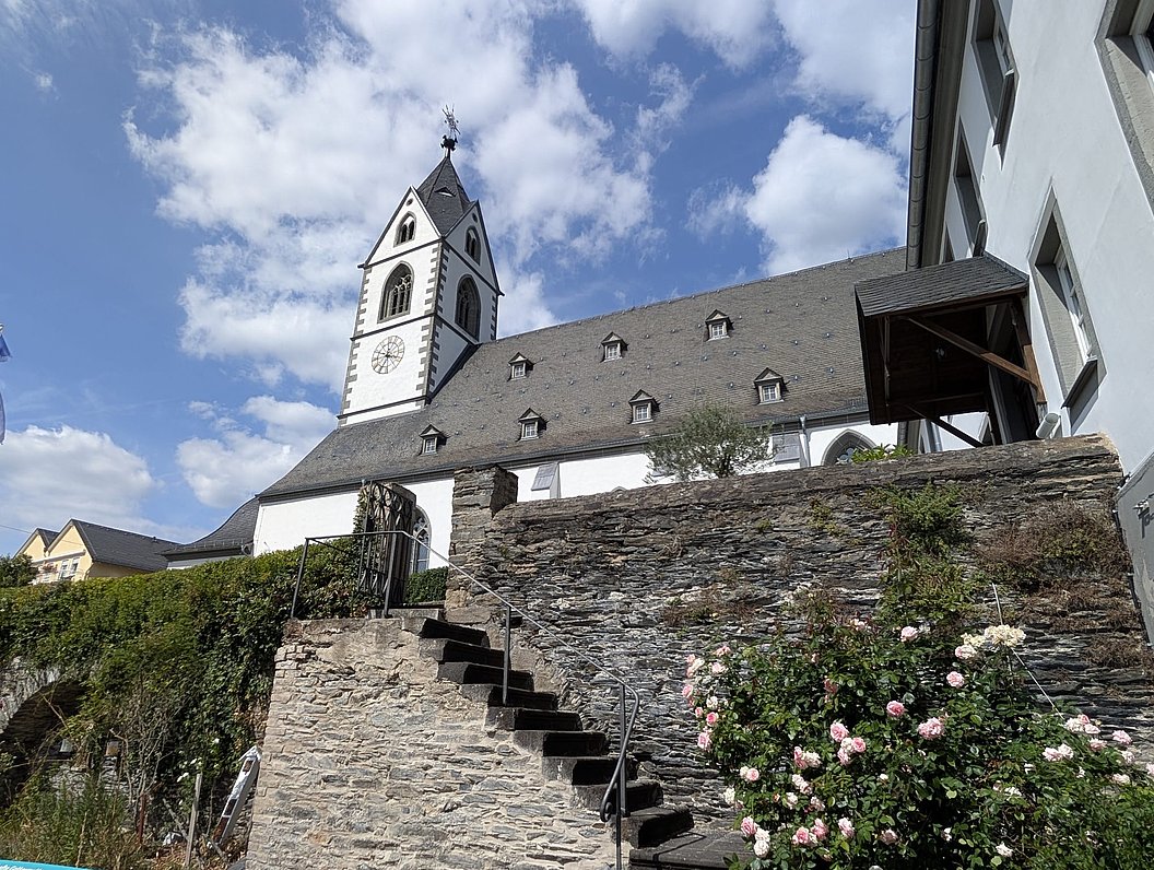 Kirchturm mit Uhr an einem historischen Gebäude hinter einer steinernen Mauer mit Treppe und blühenden Rosen