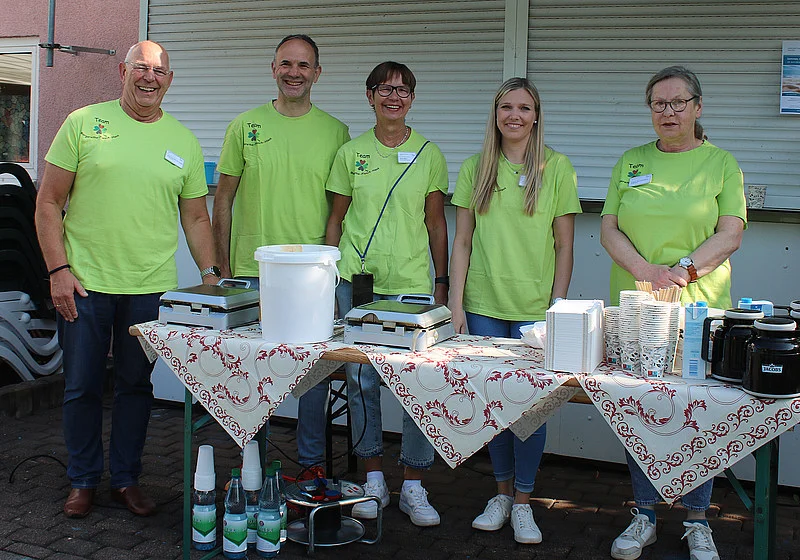 Fünf Personen in grünen T-Shirts stehen hinter einem mit Tischdecken bedeckten Tisch mit mehreren Waffeleisen, Bechern und Flaschen.