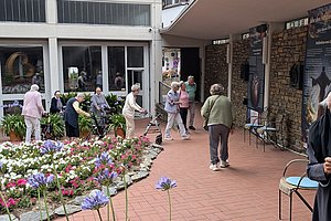 Mehrere ältere Menschen gehen und stehen auf einem gepflasterten Platz neben einem Blumenbeet vor einem Gebäude