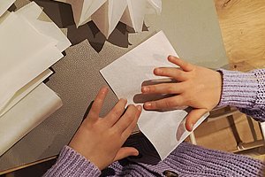 Zwei Hände falten ein weißes Papier auf einem Tisch mit weiteren Papierbögen