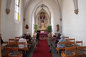 Blick in eine kleine Kirche mit Altar, mehreren älteren Menschen auf Stühlen und Rollatoren und einer stehenden Person in schwarzer Kleidung vor dem Altar