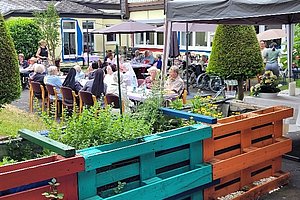 Gruppe älterer Menschen sitzt an langen Tischen im Garten eines Pflegeheims mit bunten Hochbeeten im Vordergrund