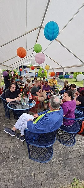 Menschen sitzen an Tischen unter einem weißen Zelt mit bunten Lampions und unterhalten sich