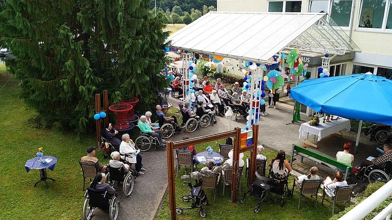 Mehrere ältere Menschen, einige im Rollstuhl, sitzen im Gartenbereich eines Gebäudes unter einem blauen Sonnenschirm und einem weißen Pavillon