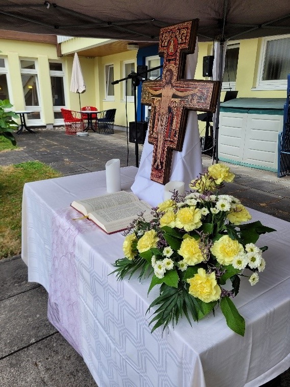 Altar mit weißem Tuch, offenem Buch, Kerze, Blumenarrangement und hölzernem Kruzifix im Innenhof eines Gebäudes